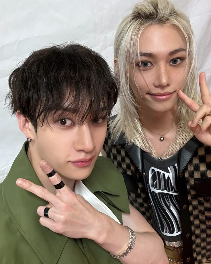 Two young men posing with peace signs, one in a green jacket and one with blonde hair in a checkered top.