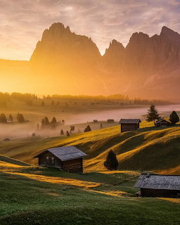 Sunrise over misty alpine valley with wooden chalets on green hillside and dramatic mountain peaks in background
