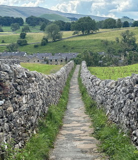 Narrow path between dry stone walls in green countryside with cottages and hills in the distance