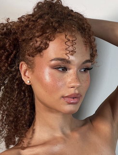 Close-up portrait of a woman with curly brown hair, glowing dewy skin, and natural makeup