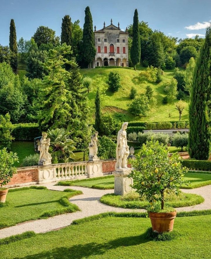 Ornate Italian garden with statues, geometric paths, cypress trees, and villa on hill