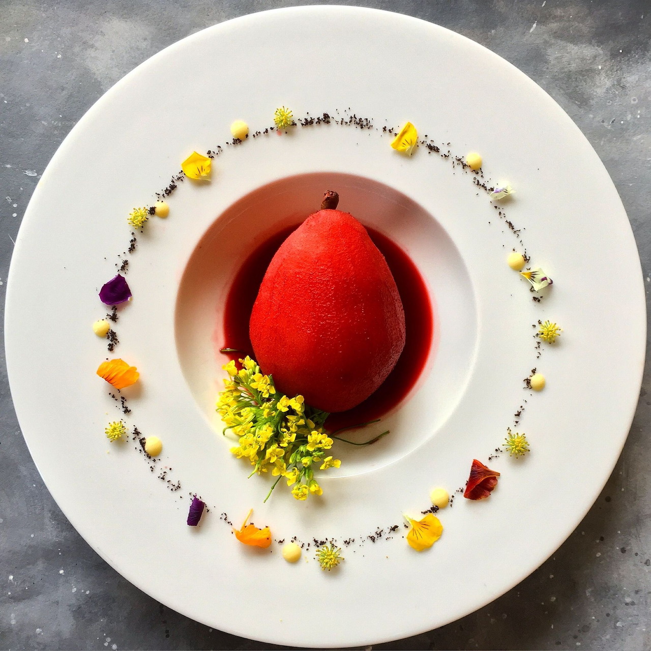 Red wine poached pear dessert with edible flower garnish on white plate
