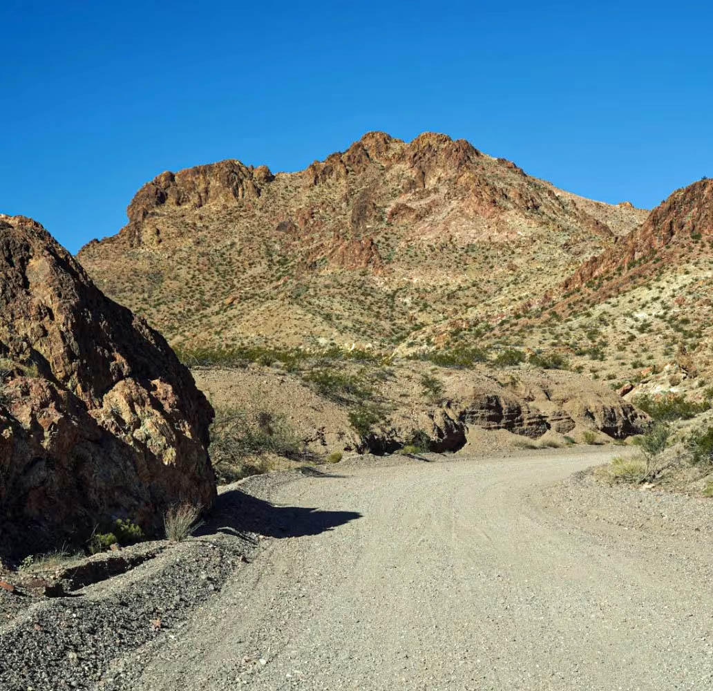 Gravel road curving through arid desert terrain with rocky mountains and blue sky