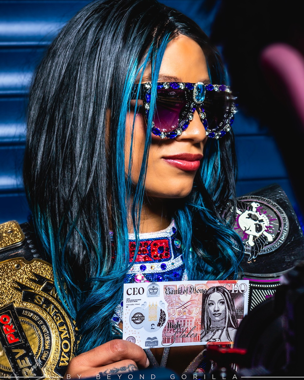 Mercedes Moné wearing jeweled sunglasses and holding a custom CEO banknote with a championship belt
