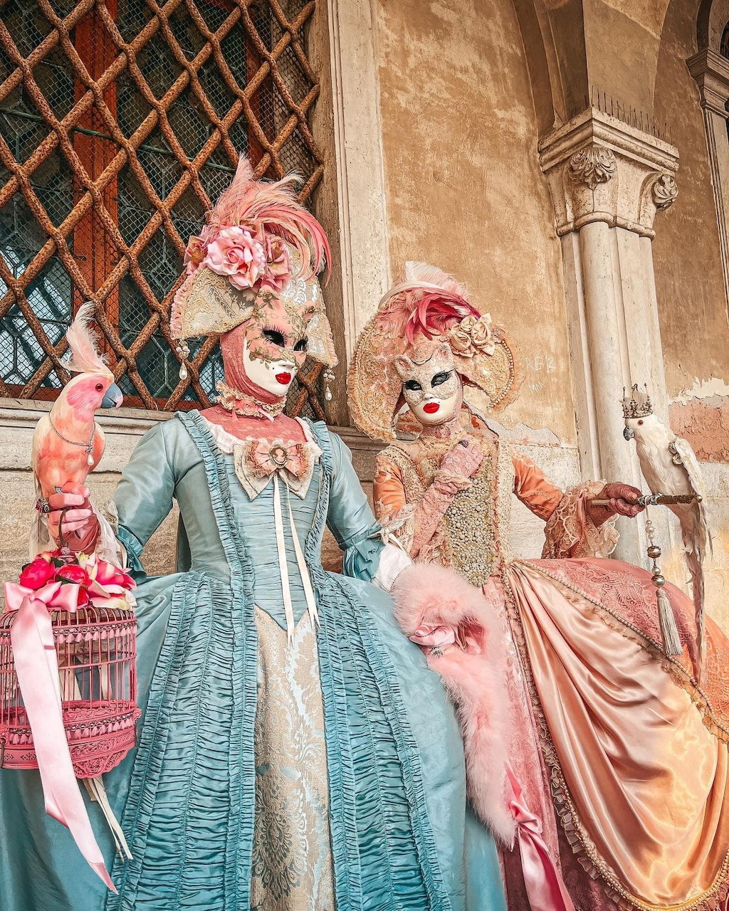 Two people in ornate Venice carnival costumes with feathered masks and period dress holding decorative birdcages