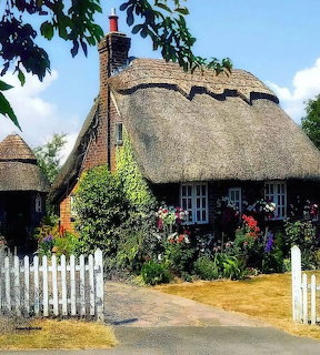 Thatched cottage with white fence and vibrant garden flowers under blue sky