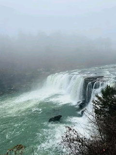 Large waterfall tumbling into a rocky gorge on a misty, overcast day with fog-covered hills in the background.