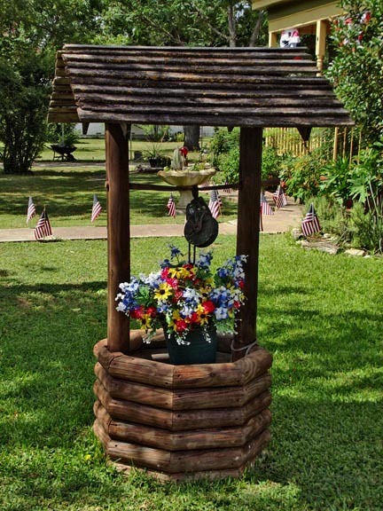 Decorative wooden wishing well planter with colorful flowers on a green lawn with American flags