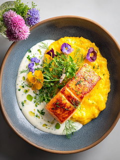 Plated salmon dish with yellow sauce, microgreens, and edible flowers in dark bowl