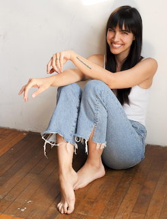 Smiling woman with long dark hair in white top and jeans sitting on wood floor