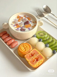 White tray with a bowl of milk dessert, moncakes, mochi, kiwi slices, blueberries, and sausages with silverware