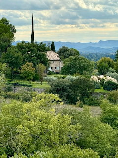 Stone cottage surrounded by green trees and shrubs with distant hills under a cloudy sky