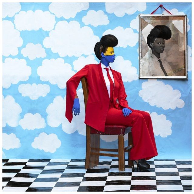 Seated woman in a red suit with painted face, cloud backdrop, checkerboard floor, and framed portrait