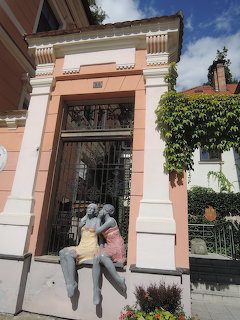 Romantic couple statue sitting on ledge before ornate pink building entrance with iron gate