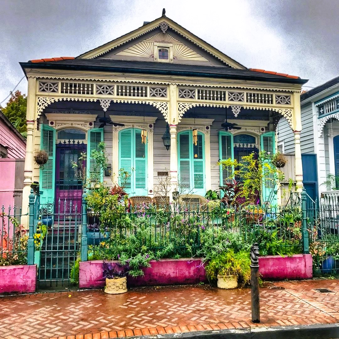 Turquoise Victorian house with decorative trim, pink planters with greenery, brick patio, cloudy sky