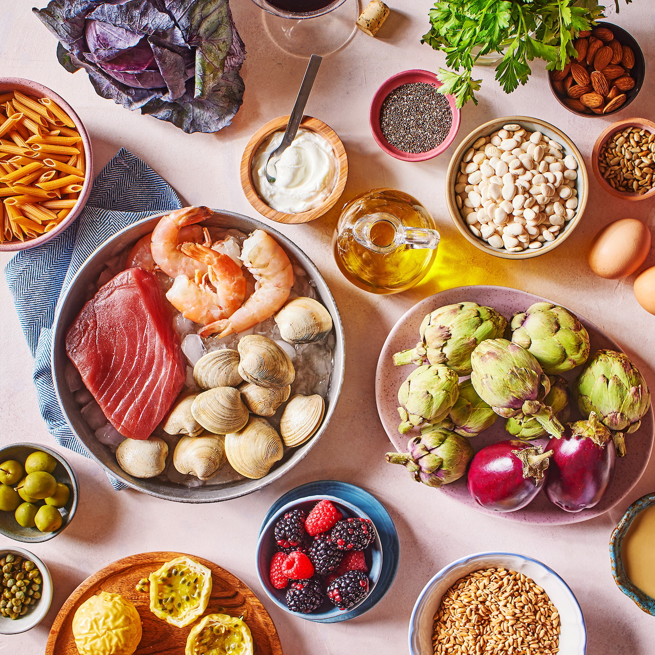 Overhead view of colorful Mediterranean diet ingredients including fish, vegetables, nuts, olive oil, and grains on white table