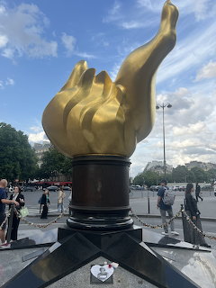 Golden Flame of Liberty sculpture on a dark pedestal in a Paris plaza with people nearby
