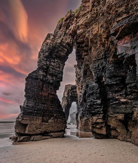Natural stone arch on beach with dramatic sunset sky and layered rock formations