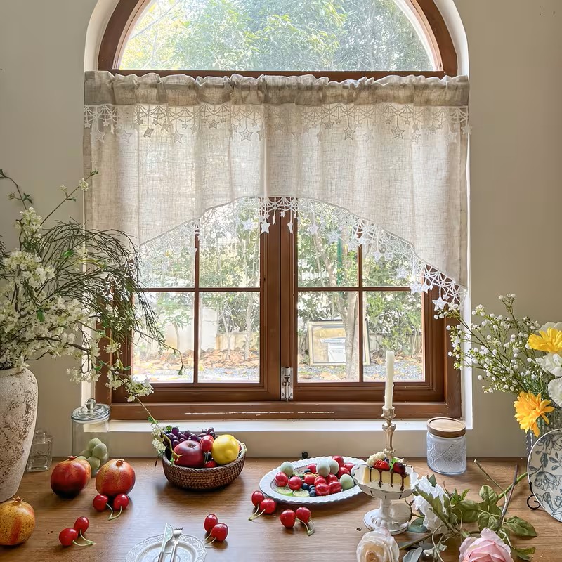 Arched window with lace curtains above a table of fruit, pastries, flowers, and teacups