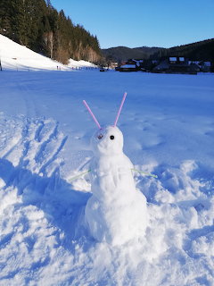 Small snowman with pink straw antennae in a snowy valley with trees and buildings in background