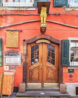 Carved wooden double doors with arched windows on coral-red building facade with green shutters and golden cat statue