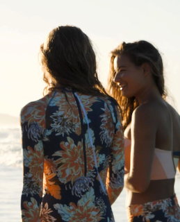 Two women seen from behind on a beach at sunset, one in floral top and the other in a bikini