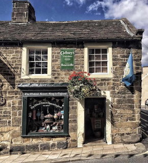 Old stone sweet shop with Cadbury sign, hanging flowers, and slate roof in an English village
