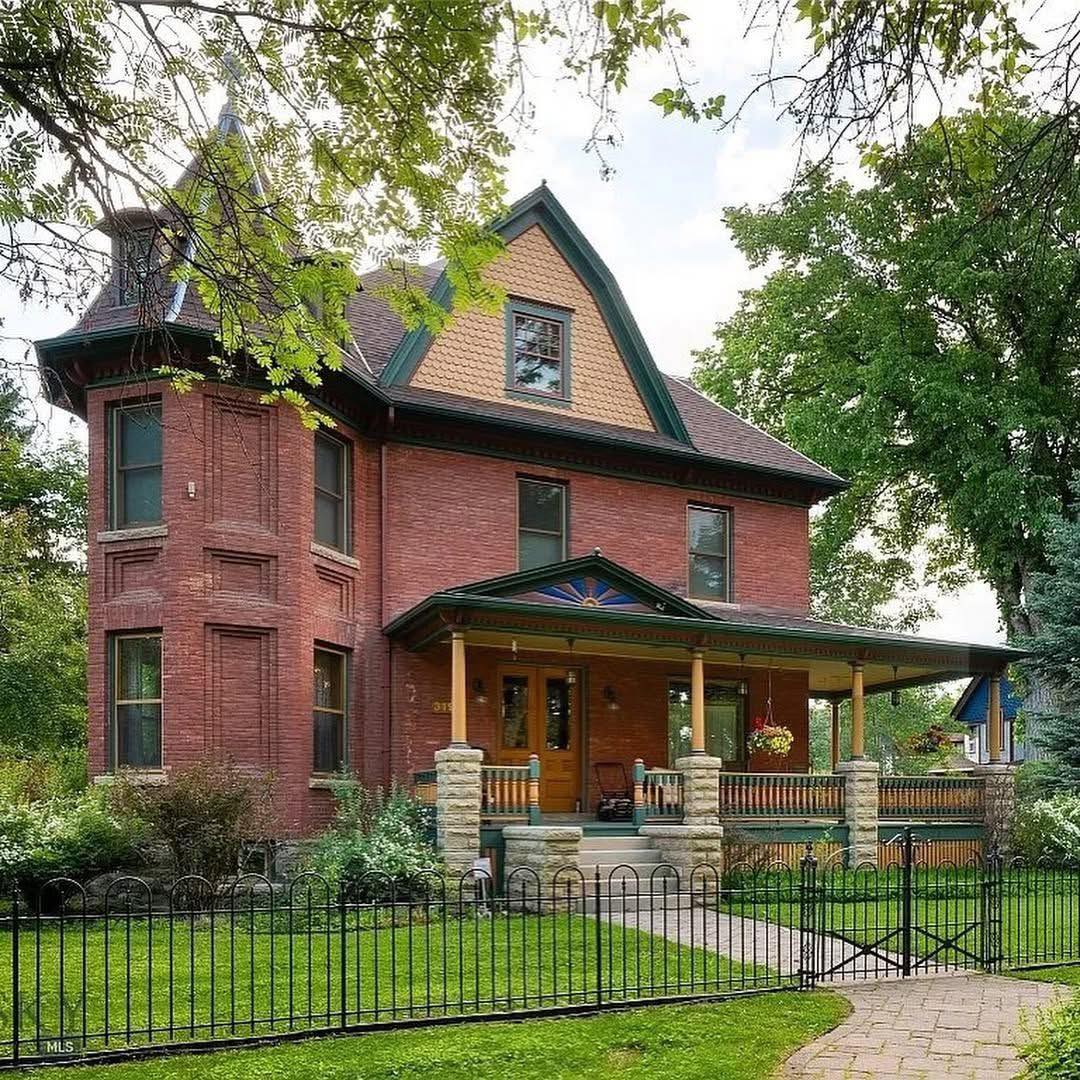 Victorian red brick house with turret and wraparound porch surrounded by green lawn and trees