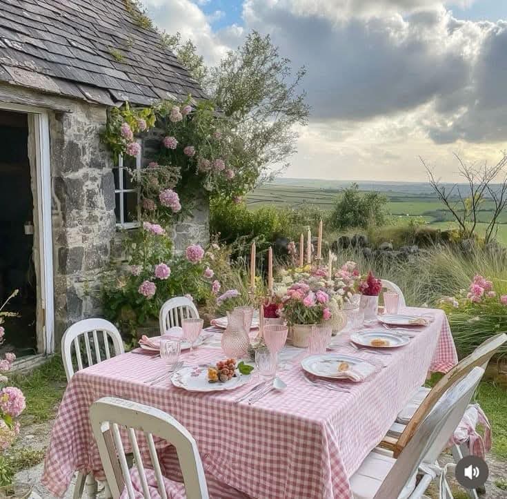 Outdoor dining table with pink tablecloth beside stone cottage, garden flowers, countryside view