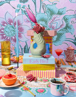 Colorful brunch table with coffee, grapefruit, pastries, books, and a flower vase against floral wallpaper