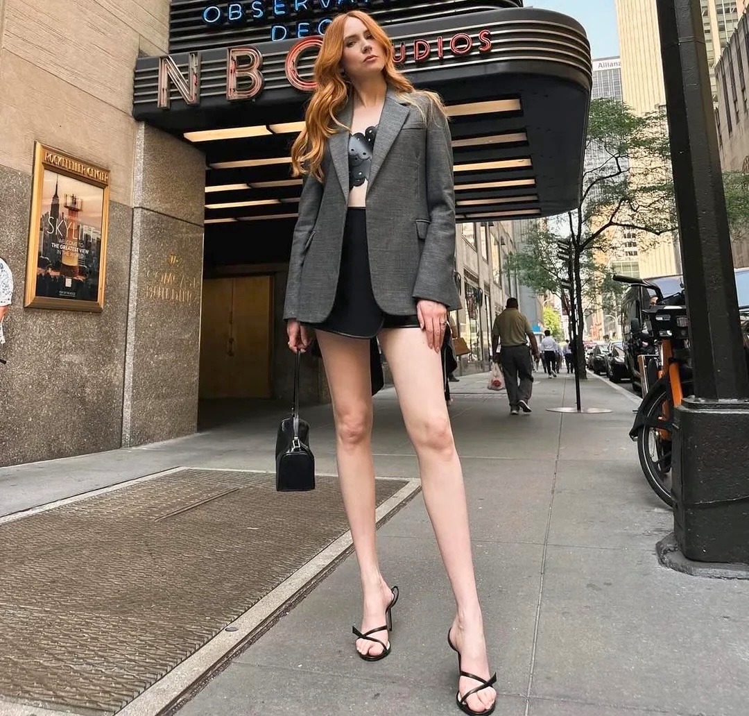 Red-haired woman in gray blazer and black mini skirt standing on NYC sidewalk near NBC building