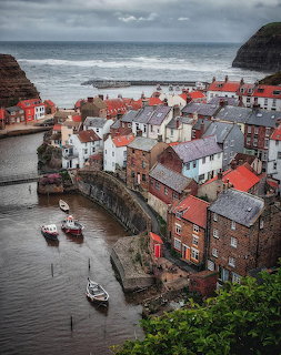 Seaside village with red-roofed houses, a narrow harbor, and small boats under a cloudy sky