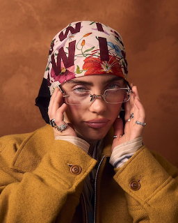Woman in floral headscarf, clear glasses, and mustard coat posing with hands near face