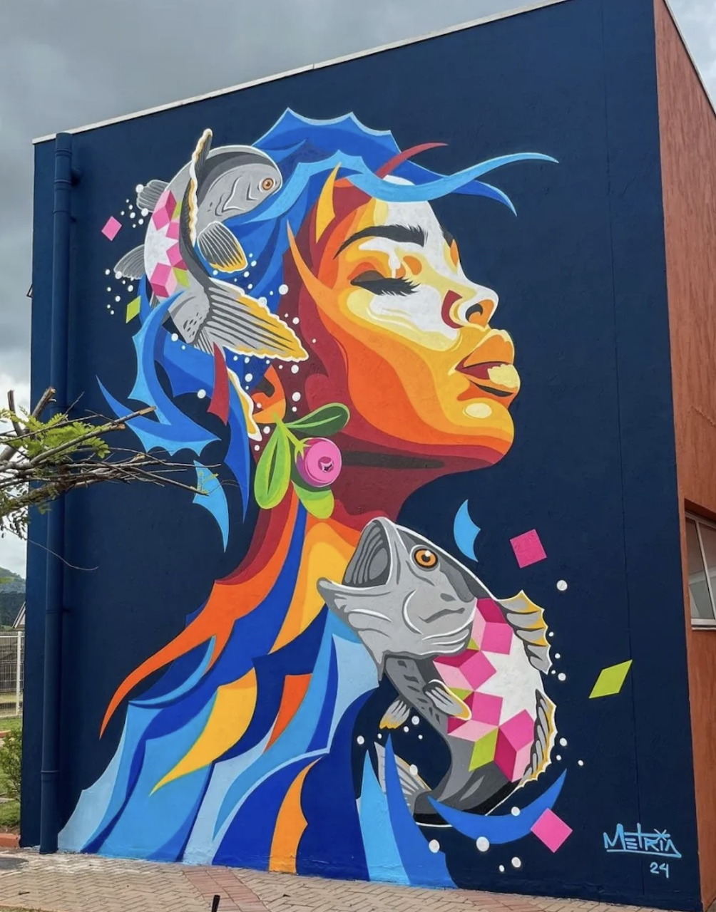Colorful street art mural of a woman with blue hair, fish, and butterfly on a dark building wall