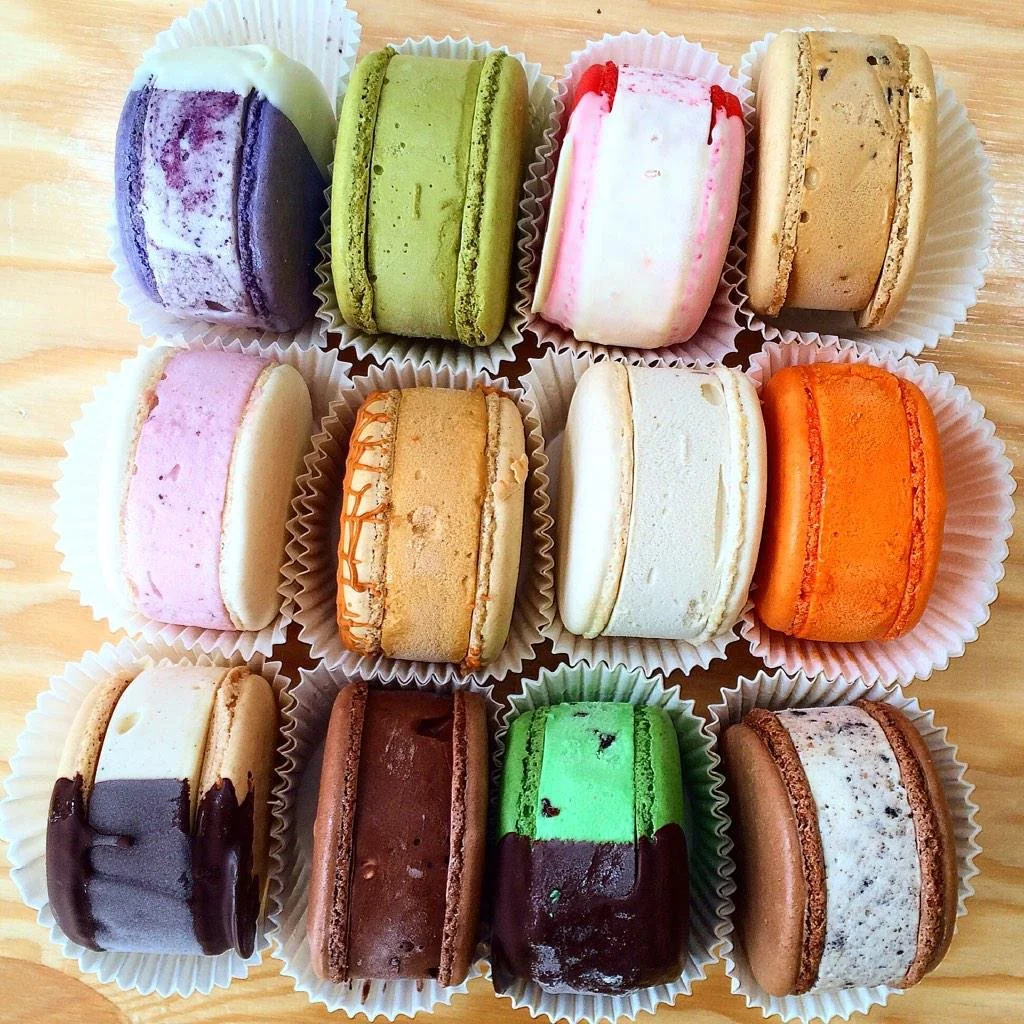 Twelve macaron ice cream sandwiches in assorted colors arranged in three neat rows on a wooden surface