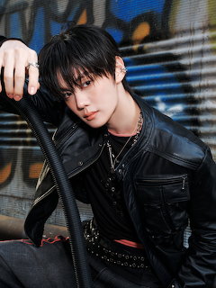 Young man with black hair in a black leather jacket and chains, posing against a graffiti-style background.