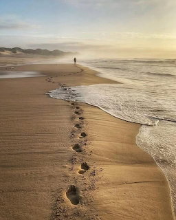 Footprints leading along a sandy beach toward a distant person with ocean waves and misty golden sky