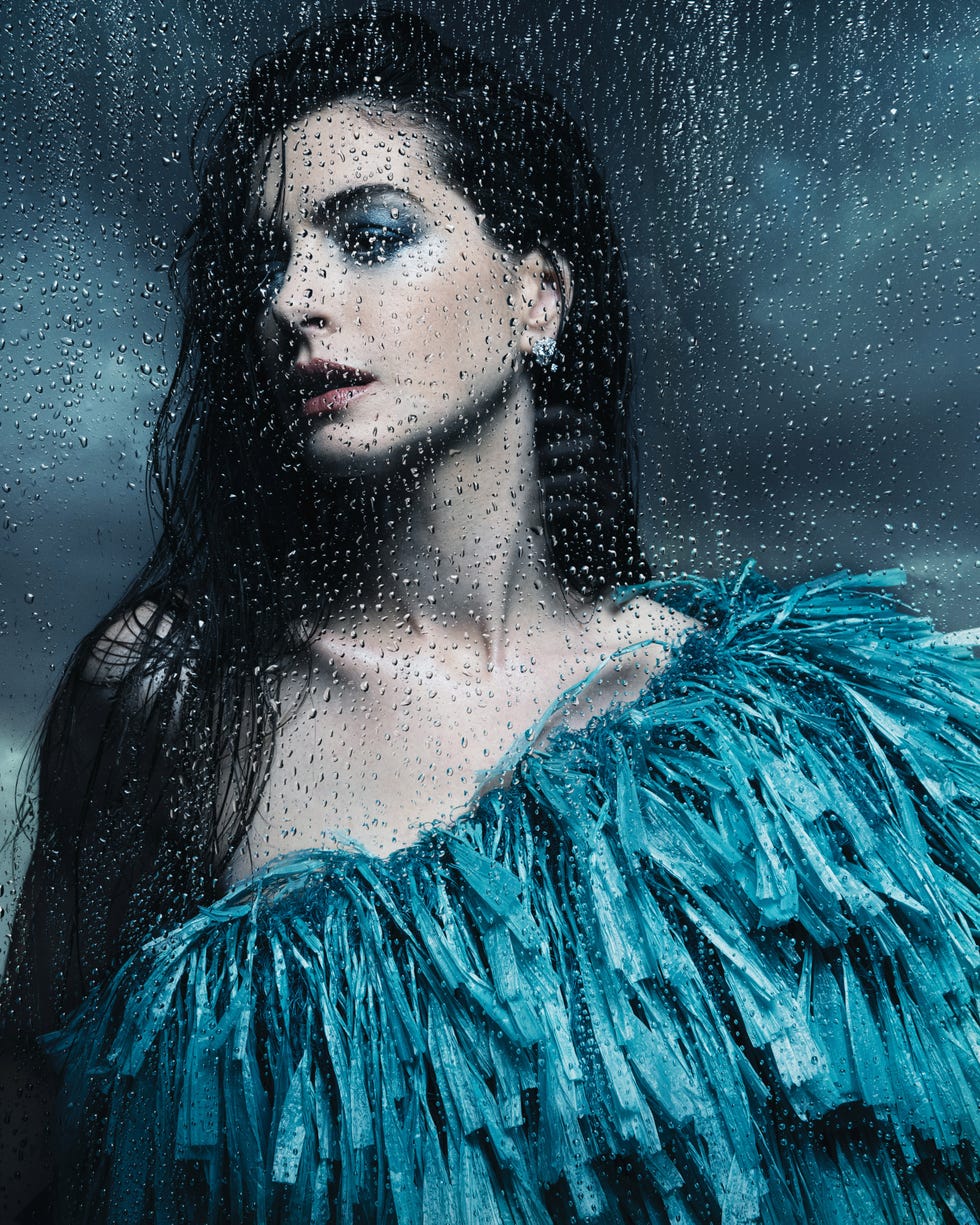 Dark-haired woman in a teal feathered dress behind rain-covered glass with blue eye makeup