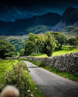 Narrow road with stone wall leading through green valley toward rugged mountains under dark sky
