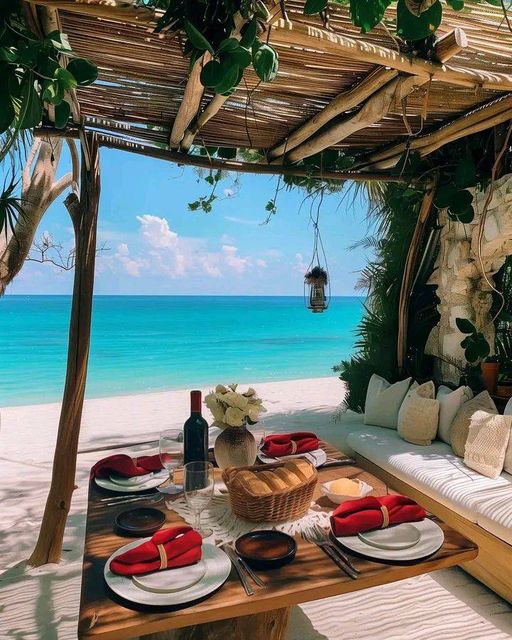 Outdoor dining table set with red napkins under a bamboo pergola overlooking a turquoise beach