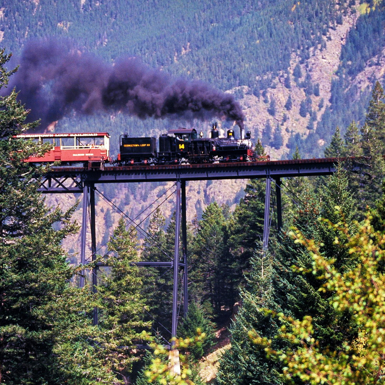 Steam train crossing tall bridge over forested mountain canyon with dark smoke plume