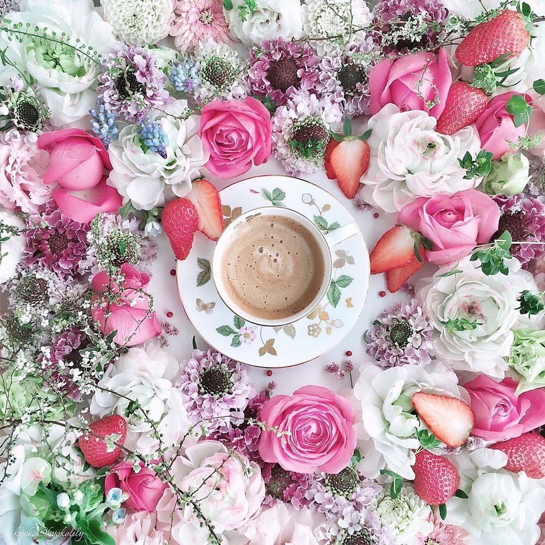 Coffee cup centered among pink roses, white flowers, and fresh strawberries in flatlay arrangement