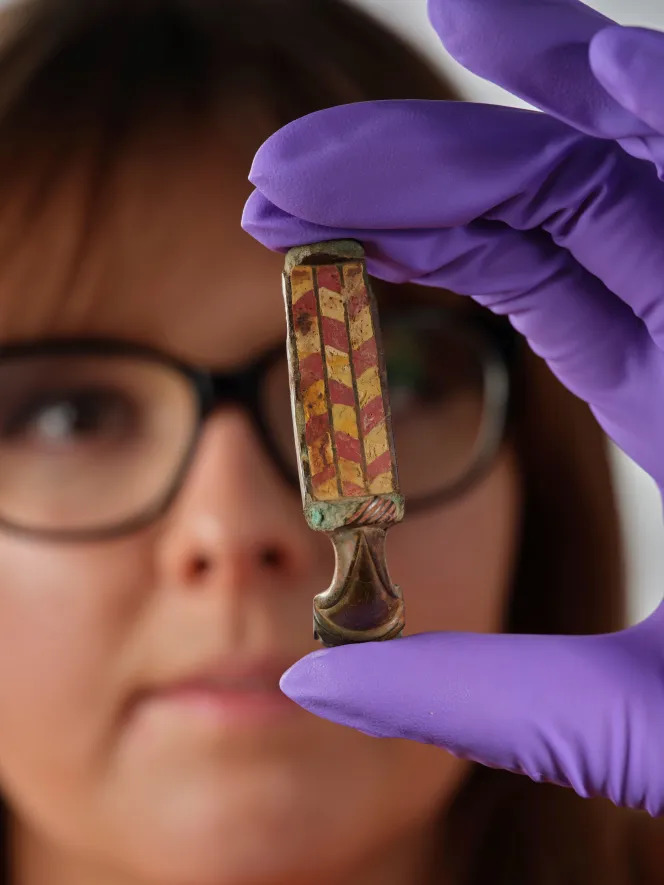 Woman in purple gloves holding ancient Roman brooch with red and gold checkered enamel pattern