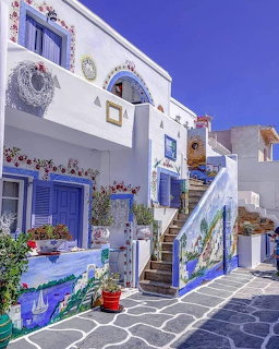 White Greek island house with blue accents, painted stairs, murals, and flower pots under blue sky