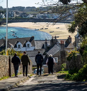 People walking down coastal path toward beach and harbour with white cottages and boats in Scilly Isles