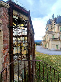 Rusty iron gate and brick pillars beside a grand chateau under a cloudy sky