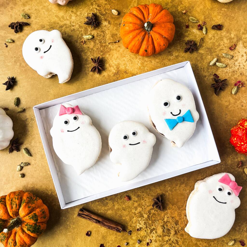 Ghost-shaped Halloween macarons with cute faces in a white box, surrounded by pumpkins and spices