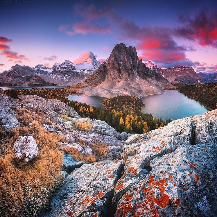 Mount Assiniboine peak rising above a turquoise glacial lake at sunset with rocky foreground and colorful clouds