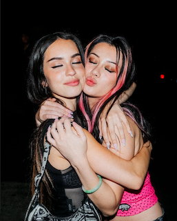 Two young women hugging and posing together at night, one making a kiss face with pink hair highlights