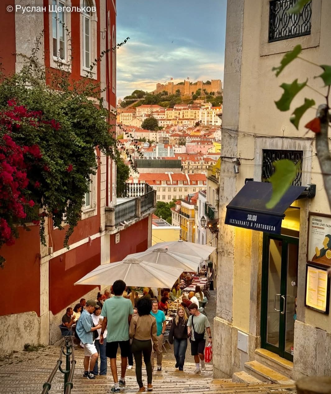 Narrow Lisbon street with tourists, colorful buildings on hillside and castle in background at golden hour